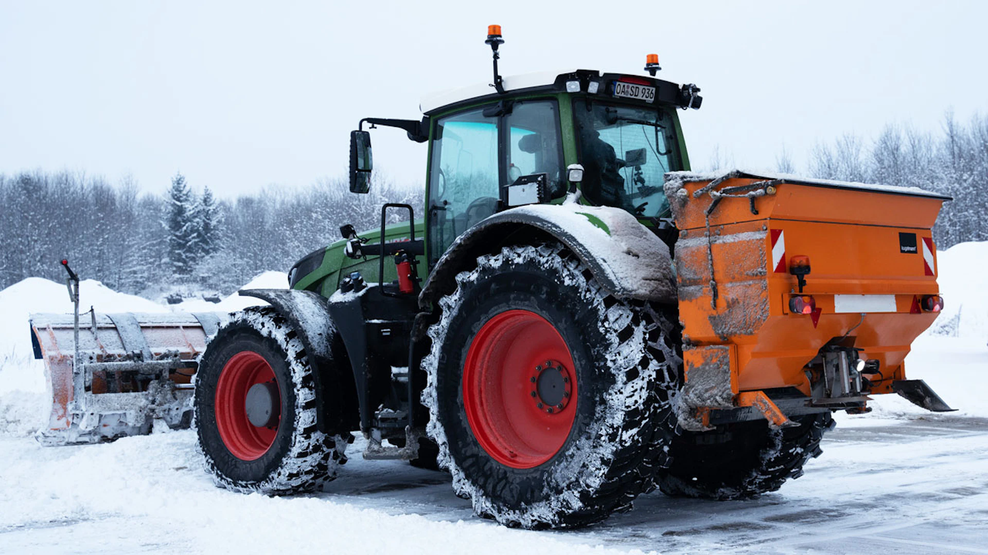 Traktor mit einem Streuapparat hinten drauf, vorne ist eine große Schneeschaufel angebracht.