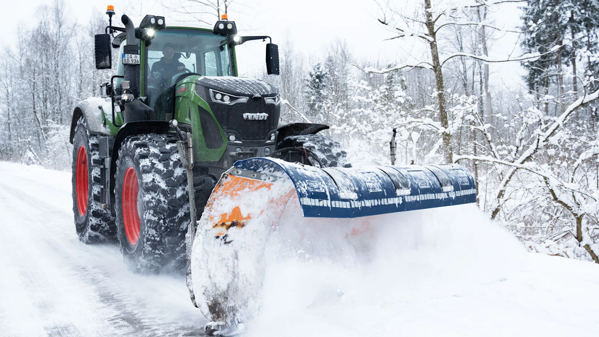Traktor mit einer großen Schneeschaufel der an einem Waldstück Schnee wegschiebt