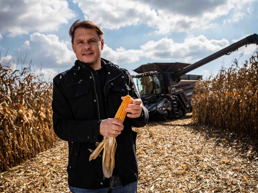 Josef Götz steht mit einem Maiskolben in der Hand auf dem Feld. Hinter ihm ist ein Fendt-Mähdrescher zu sehen.